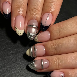 Può includere: Unghie curate con base nude e design vari tra cui punte alla francese nere, fasce orizzontali metalliche argentate, piccoli borchie argentate e punte color crema con pois neri. Le unghie hanno una forma a mandorla arrotondata.