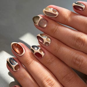 Puede incluir: Uñas de forma ovalada con una base de color crema y diseños de espacio negativo. Cada uña muestra esmalte brillante con efecto ojo de gato en tonos rojo oscuro, marrón, gris y dorado, enmarcado por bordes suaves y elevados de color crema.