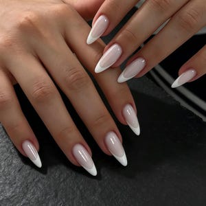 Op de afbeelding: Lange, amandelvormige vingernagels met een klassiek Frans manicureontwerp. De nagels hebben een transparante, lichtroze basiskleur met strakke, dekkende witte uiteinden die de gebogen rand van de nagel volgen voor een verzorgde uitstraling.