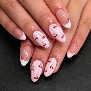 Può includere: Unghie a forma di mandorla con un mix di design. Alcune unghie presentano una classica manicure francese con punte rosa chiaro, mentre altre mostrano un motivo a righe verticali rosa e bianco decorato con piccole illustrazioni di ciliegie.