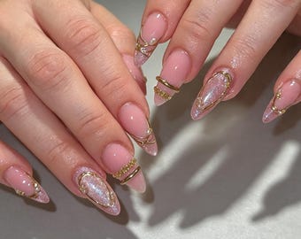 May include: Almond-shaped nails featuring a soft pink base with a shimmering, iridescent finish. The nail art includes delicate gold wire accents, thin metallic rings, and small white pearls placed along the tips and across the nail beds.