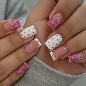 Può includere: Unghie di forma quadrata con un mix di design, tra cui smalto bianco a pois rossi, punte rosa e basi trasparenti. Diverse unghie sono decorate con accenti floreali rosa tridimensionali e piccoli strass argentati incastonati.