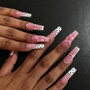 Può includere: Unghie artificiali lunghe di forma quadrata con stile french manicure su base rosa traslucida. Alcune punte sono bianche a pois neri, mentre altre presentano decorazioni floreali rosa tridimensionali con piccole perline dorate al centro.
