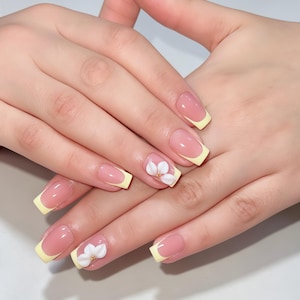 May include: A set of square-shaped fingernails featuring a classic French manicure style with pale yellow tips. Two of the nails are accented with a three-dimensional white flower design, each featuring a small gold-coloured centre detail.