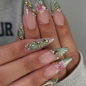 Puede incluir: Uñas en forma de almendra con base nude y puntas verde salvia. El diseño incluye flores rosas en 3D, detalles de filigrana dorada, pequeñas perlas blancas y crestas verdes texturizadas, creando una apariencia superpuesta y ornamentada.