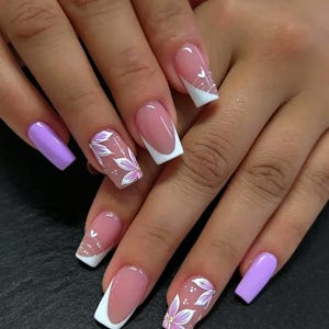Può includere: Unghie di forma quadrata con un mix di smalto color viola chiaro e uno stile french manicure con punte bianche. Due unghie sono decorate con disegni floreali dipinti a mano in bianco e viola, accentuati da piccoli puntini bianchi su base trasparente.