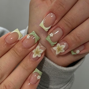 May include: Square-shaped nails featuring a nude base with sage green French tips. The design includes three-dimensional white floral accents with gold centers on select nails, creating a textured look across the manicured fingernails.