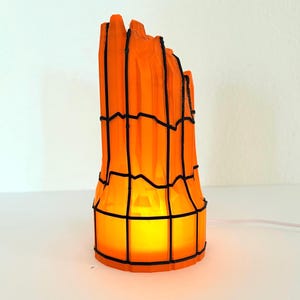 以下が含まれることがあります： ユニークな建築デザインのオレンジと黒の装飾ランプ。 ランプはケージのような構造で、内側から暖かい黄色の光が発せられています。 白いコンセントに差し込まれています。