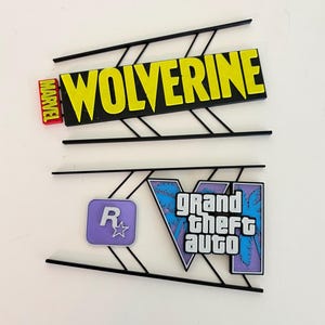 May include: Two decorative wall signs featuring black geometric frames. The top sign displays the yellow text WOLVERINE with a red MARVEL logo. The bottom sign features a purple Rockstar Games logo and a blue and purple Grand Theft Auto VI logo.