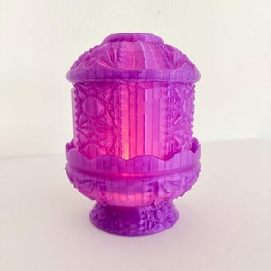 May include: A vibrant purple glass candle holder featuring a faceted, geometric pressed pattern across its cylindrical body, domed lid, and scalloped base. The translucent material catches light, highlighting the intricate star and diamond textures.