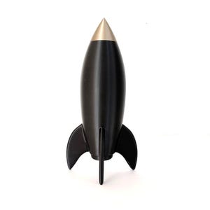 Pode incluir: Um objeto em forma de foguete com um corpo preto fosco e um cone de nariz cônico de cor dourada metálica lisa. A estrutura apresenta três barbatanas curvas que se estendem da base, proporcionando uma postura estável de tripé para o foguete.
