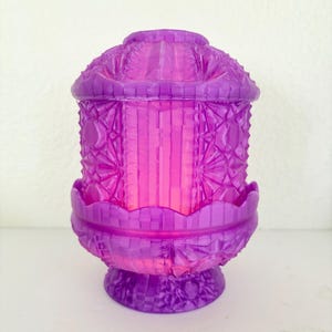 May include: A decorative purple lantern with a detailed, geometric design. The lantern has a domed top, a cylindrical body, and a scalloped base. The interior glows with a soft pink light, creating a warm ambiance.