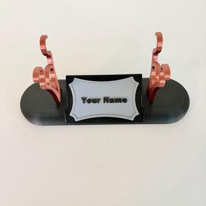 Puede incluir: Un soporte de exhibici&oacute;n ovalado negro con dos soportes verticales de color cobre met&aacute;lico con recortes decorativos curvos. Una placa rectangular gris claro con borde festoneado muestra el texto negro Your Name en el centro.