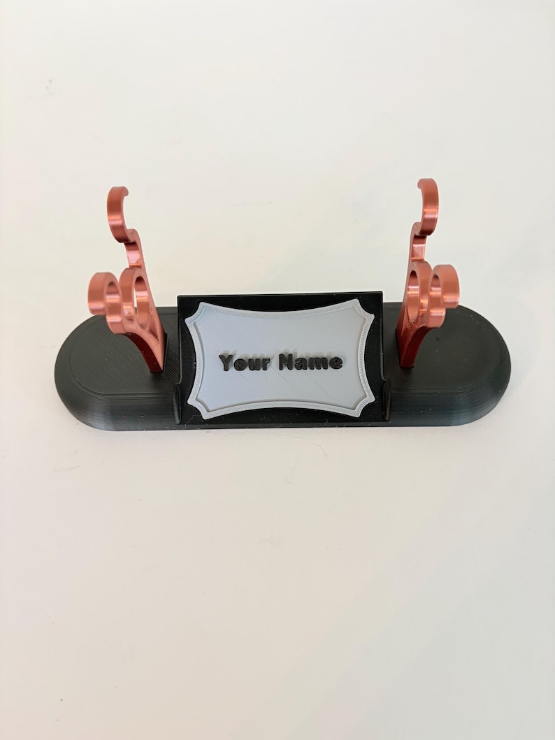 Puede incluir: Un soporte de exhibici&oacute;n ovalado negro con dos soportes verticales de color cobre met&aacute;lico con recortes decorativos curvos. Una placa rectangular gris claro con borde festoneado muestra el texto negro Your Name en el centro.