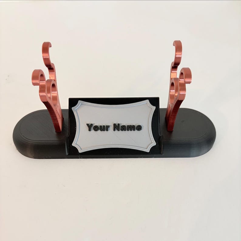 Puede incluir: Un soporte de base ovalada negra con dos soportes met&aacute;licos de color cobre con ganchos curvos. Una placa rectangular central con borde festoneado est&aacute; fijada en la parte frontal, mostrando el texto negro en relieve Your Name.