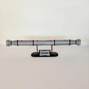 Puede incluir: Una empu&ntilde;adura de sable de luz plateada de doble hoja con detalles negros y botones rojos, montada horizontalmente sobre un soporte negro. El soporte incluye una placa plateada con el texto Your Name centrado en la base frontal.