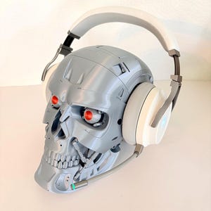 May include: A silver robotic skull with glowing red eyes wearing white and silver over-ear headphones. The headphones feature a flexible microphone boom arm extending from the side, with a small green light indicator at the tip of the microphone.