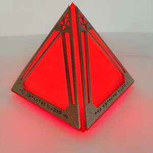 Peut inclure: Une lampe en forme de pyramide avec un cadre en bronze métallique texturé et des panneaux lumineux rouge vif. La base du cadre présente des symboles gravés complexes sur chaque côté, et la structure émet une lumière rouge éclatante.