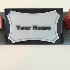 Puede incluir: Un soporte para placa de identificaci&oacute;n de escritorio negro con una placa rectangular gris claro de bordes festoneados decorativos. El texto "Your Name" est&aacute; en relieve con letras negras en el centro de la placa. La base es negra con detalles rojos.