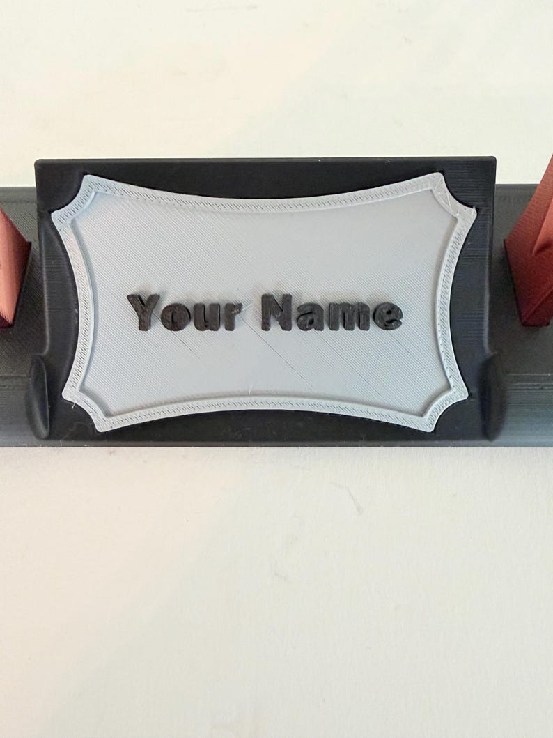 Puede incluir: Un soporte para placa de identificaci&oacute;n de escritorio negro con una placa rectangular gris claro de bordes festoneados decorativos. El texto "Your Name" est&aacute; en relieve con letras negras en el centro de la placa. La base es negra con detalles rojos.