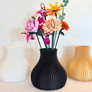 May include: A black ribbed vase displays a vibrant arrangement of artificial flowers. Two other vases, one white and one yellow, are positioned nearby. The flowers feature pink, orange, and yellow petals, with green stems and foliage.