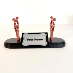 Puede incluir: Un soporte de exhibici&oacute;n horizontal negro con dos soportes de color cobre con ganchos curvos. Una placa rectangular plateada con un borde decorativo est&aacute; fijada en la parte frontal, mostrando el texto negro Your Name.