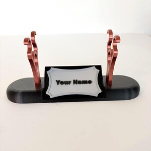 Puede incluir: Un soporte de base ovalada negra con dos soportes met&aacute;licos de color cobre con ganchos curvos y una placa central rectangular plateada con borde decorativo. La placa muestra el texto negro en relieve Your Name.