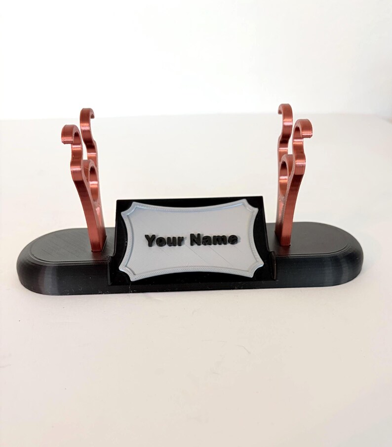 Puede incluir: Un soporte de base ovalada negra con dos soportes met&aacute;licos de color cobre con ganchos curvos y una placa central rectangular plateada con borde decorativo. La placa muestra el texto negro en relieve Your Name.