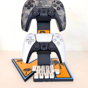 May include: A black vertical gaming controller stand holding a gray camouflage controller on top and a white controller on the bottom. The base features an orange and blue design with palm tree silhouettes and white 3D text reading Grand Theft Auto.
