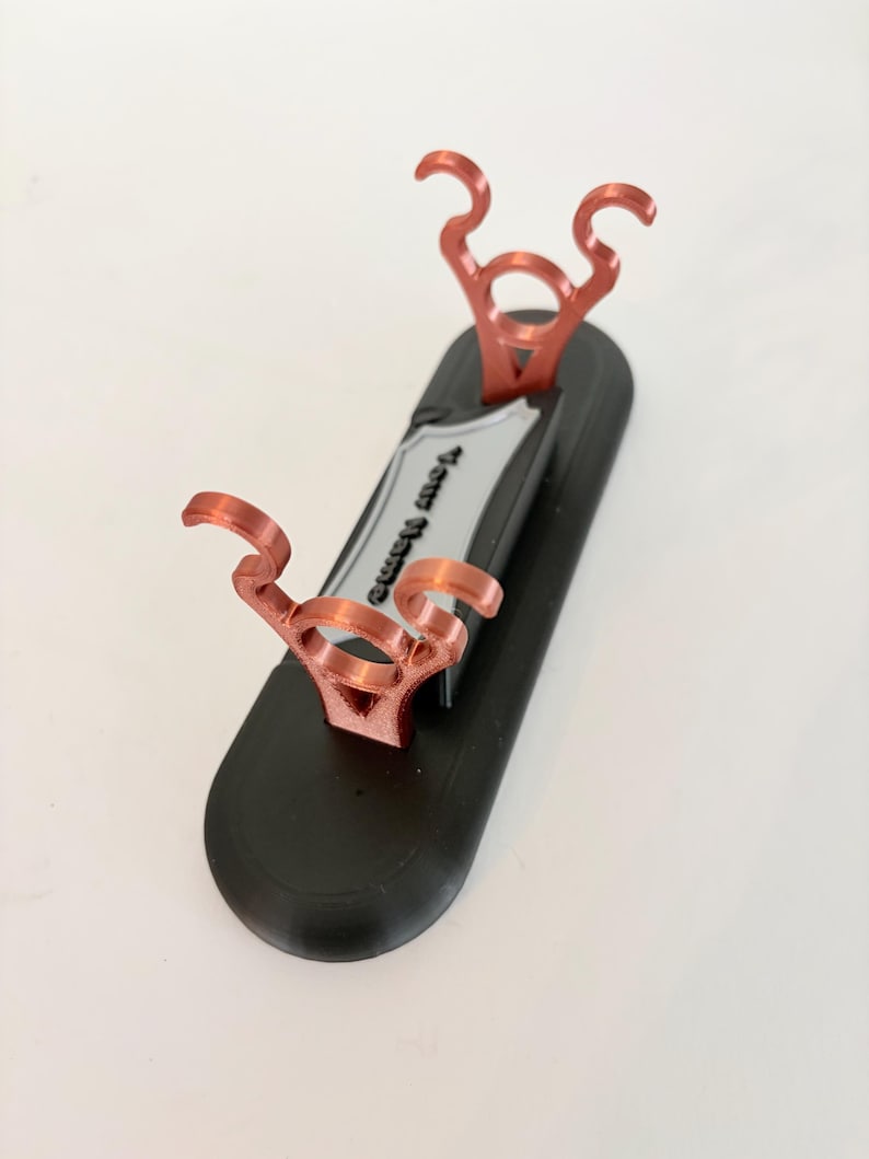 Puede incluir: Un soporte de base ovalada negra con dos soportes de color cobre met&aacute;lico con partes superiores curvas. Una placa de identificaci&oacute;n rectangular de color plateado se sit&uacute;a entre los soportes, mostrando el texto negro en relieve "Your Name".