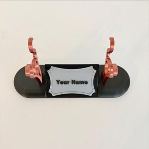 Puede incluir: Un soporte de exhibici&oacute;n ovalado negro con dos soportes verticales de color cobre met&aacute;lico y una placa rectangular central de color gris claro con un borde decorativo. La placa tiene el texto negro Your Name en relieve en su parte frontal.