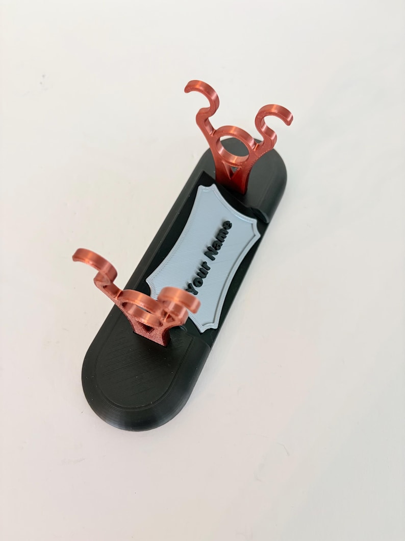 Puede incluir: Un soporte de exhibici&oacute;n ovalado negro con dos soportes verticales de color cobre met&aacute;lico con partes superiores curvas. Una placa gris claro en forma de escudo est&aacute; centrada en la base, mostrando el texto negro "Your Name".