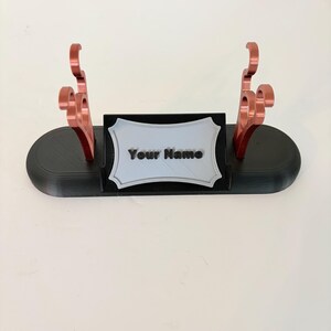 Puede incluir: Un soporte de exhibici&oacute;n ovalado negro con dos soportes verticales de color cobre con ganchos curvos. Una placa rectangular central con borde festoneado es de color gris claro y muestra el texto negro en relieve "Your Name" al frente.