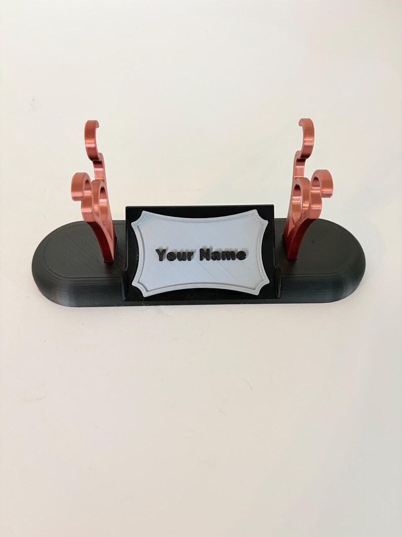 Puede incluir: Un soporte de exhibici&oacute;n ovalado negro con dos soportes verticales de color cobre con ganchos curvos. Una placa rectangular central con borde festoneado es de color gris claro y muestra el texto negro en relieve "Your Name" al frente.
