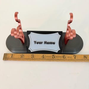 Puede incluir: Un soporte de exhibici&oacute;n negro con dos soportes met&aacute;licos de color cobre y una placa central gris claro con el texto "Your Name" en negro. Una regla de madera colocada delante muestra que la base mide aproximadamente 20 cm de largo.