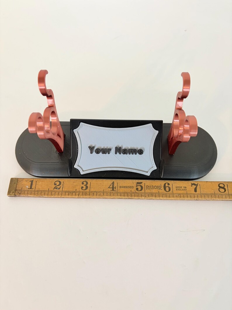 Puede incluir: Un soporte de exhibici&oacute;n negro con dos soportes met&aacute;licos de color cobre y una placa central gris claro con el texto "Your Name" en negro. Una regla de madera colocada delante muestra que la base mide aproximadamente 20 cm de largo.