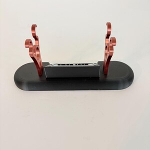 Puede incluir: Un soporte de base ovalada negra con dos soportes met&aacute;licos de color cobre con ganchos curvos en la parte superior. Un peque&ntilde;o bloque rectangular negro con un borde superior plateado est&aacute; colocado centralmente entre los dos soportes.