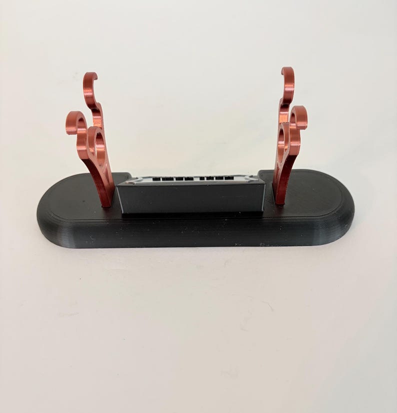 Puede incluir: Un soporte de base ovalada negra con dos soportes met&aacute;licos de color cobre con ganchos curvos en la parte superior. Un peque&ntilde;o bloque rectangular negro con un borde superior plateado est&aacute; colocado centralmente entre los dos soportes.