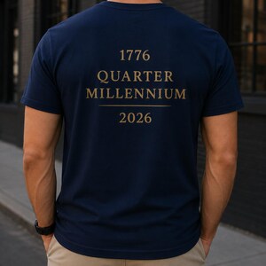 Peut inclure: Un t-shirt bleu marine à manches courtes avec un texte doré au dos indiquant 1776 QUARTER MILLENNIUM 2026, accompagné d'une ligne horizontale séparant les mots de l'année. Le t-shirt est porté avec un pantalon kaki et une montre noire.