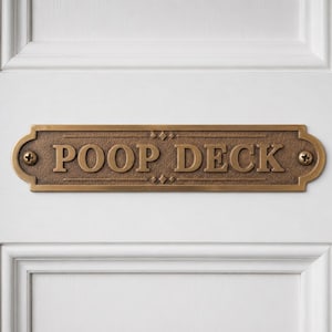 May include: A rectangular brass plaque with rounded, decorative ends, featuring the embossed text POOP DECK in capital letters. The plaque has a textured, dark bronze-colored finish and is secured to a white paneled surface with two screws.