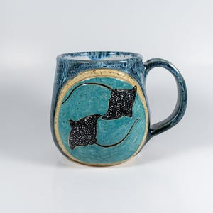 May include: A ceramic mug with a rounded body and a handle, featuring a speckled blue glaze. The center displays a circular medallion with a turquoise background and two black stingrays with white spots swimming in a circular motion.