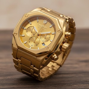 Könnte beinhalten: Eine goldfarbene Invicta Chronographenuhr mit passendem goldenem Armband. Das Zifferblatt der Uhr verfügt über mehrere Hilfszifferblätter und ein Datumsfenster. Die Uhr hat ein rechteckiges Gehäuse mit einer strukturierten Lünette und einem goldfarbenen Zifferblatt.