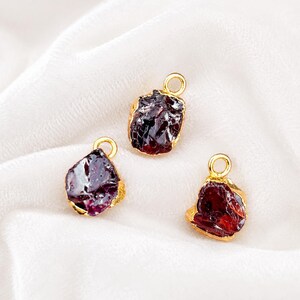 May include: Three raw garnet gemstone pendants with gold-colored metal edging and loops. The gemstones are a deep red-purple color, and the pendants are displayed on a white, textured fabric.
