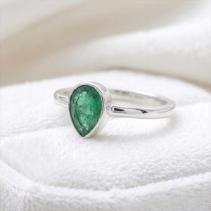 May include: A silver ring with a pear-cut, vivid green emerald gemstone. The emerald is set in a bezel setting, and the ring band is smooth and polished. The ring is presented on a white, textured surface.