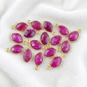 May include: Marquise-cut ruby gemstones with gold-coloured bezels and loops. The gemstones are a deep magenta colour and are arranged on a white surface. These are likely jewellery-making components.