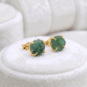 Puede incluir: Un par de pendientes de botón con piedras de esmeralda verde en bruto y texturizadas, engastadas en garras de metal dorado. Cada pendiente presenta una piedra preciosa circular de corte irregular sujeta por cuatro pequeñas garras metálicas.