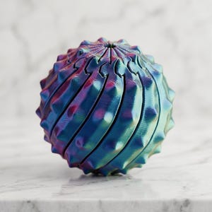 May include: A spherical object with a textured, spiky surface featuring a color-shifting iridescent finish that transitions between shades of deep blue, purple, and teal. The structure is composed of multiple interlocking segments.