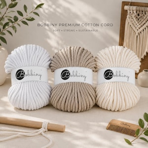 May include: Three spools of Bobbiny premium cotton cord in white, beige, and tan. Each spool has a white label with the Bobbiny logo. The text "Soft, Strong, Sustainable" is above the spools. A wooden dowel and comb are in the foreground.