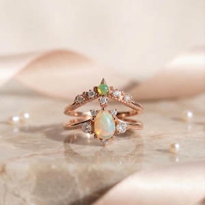 May include: A rose gold ring set featuring two bands. The lower band has a pear-shaped opal centre stone flanked by small round clear stones. The upper band has a small round opal at the peak, accented with additional round clear stones.