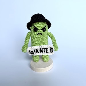 May include: A light green crocheted pickle figure wearing a black hat, featuring an angry facial expression with black eyes and a stitched mouth. The figure holds a white crocheted sign with the black embroidered text WANTED, standing on a round base.